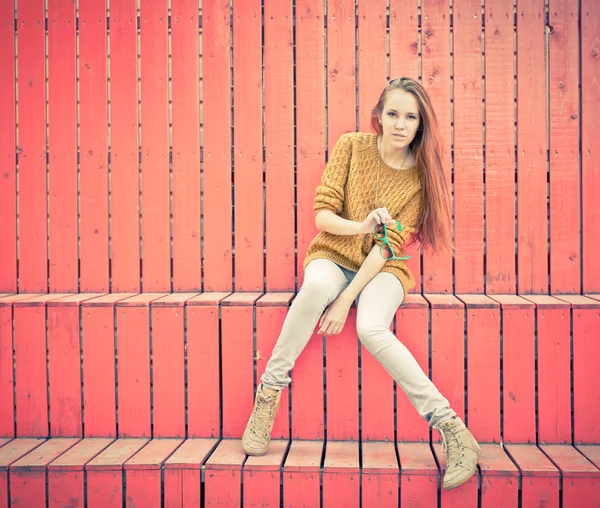 Beautiful redheaded girl  in jeans sits near wall of red wooden planks — Stock Photo, Image