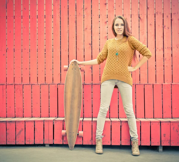 Hermosa chica pelirroja sosteniendo un longboard de pie cerca de la pared roja — Foto de Stock