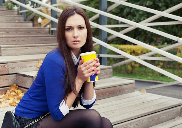 Beautiful brunette sitting on wooden steps with a glass and looking at the camera — Stock Photo, Image