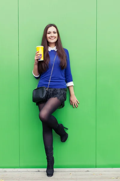 Красивая брюнетка, стоящая со стаканом в руке у зеленой стены и улыбающаяся в камеру — стоковое фото