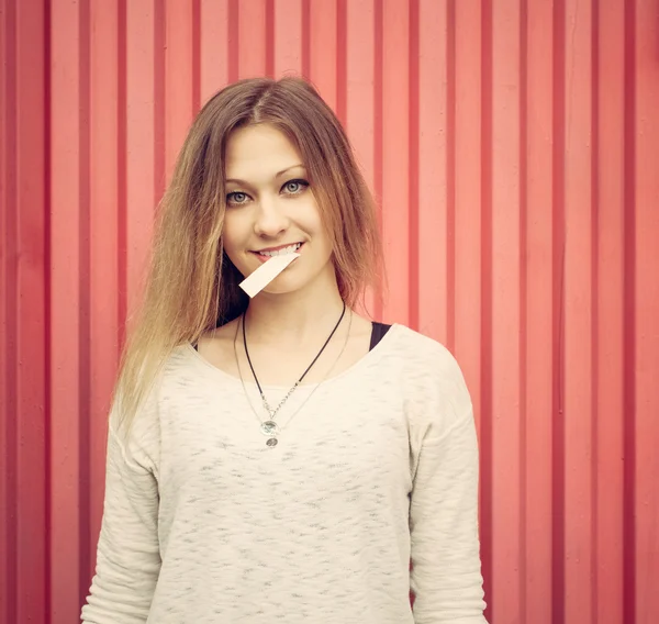 Hermosa joven rubia con goma de mascar en los labios posando cerca de la pared roja. Exterior —  Fotos de Stock