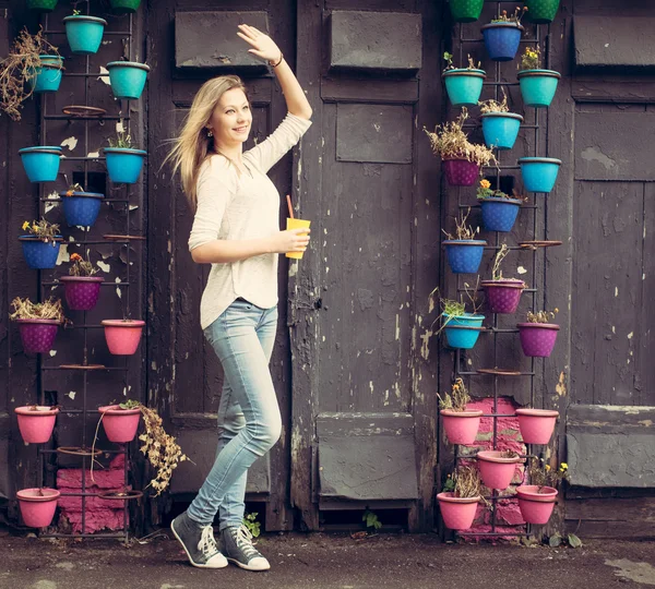 Mädchen in Jeans und Turnschuhen mit einer gelben Tasse winkt an der Wand mit Blumen. Außenbereich — Stockfoto