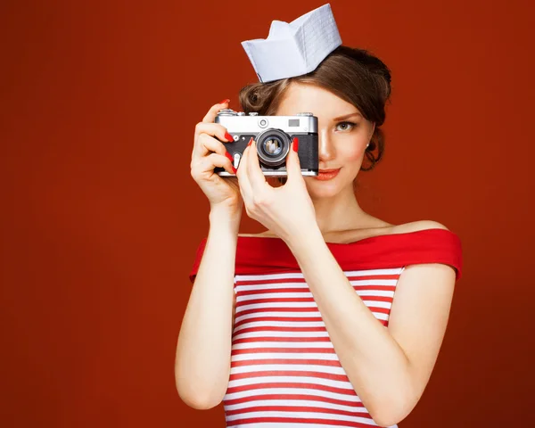 Bella pin-up ragazza in possesso di una fotocamera vintage e lo dirige direttamente alla fotocamera. sfondo rosso, primo piano — Foto Stock