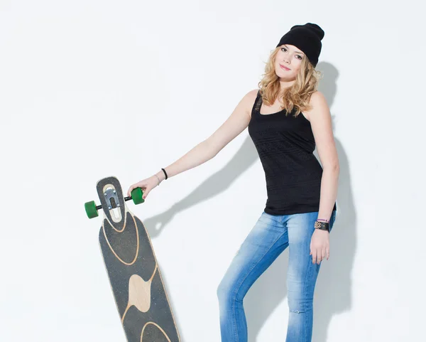 Mooi modieuze heldere jonge meisje in jeans, een zwart T-shirt en hoed poseren met een koele longboard in de handen van in de buurt van een witte muur. Binnen. Studio. — Stockfoto