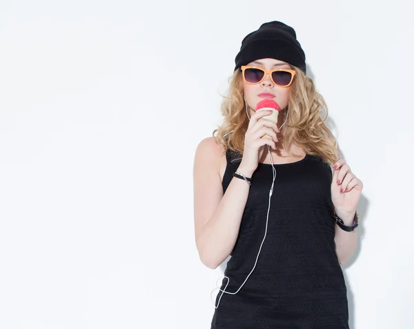 Schöne junge blonde sexy Frauen mit Sonnenbrille, schwarzem T-Shirt und Hut, die Musik hören und Eis essen. Nahaufnahme. Hintergrund Studio-Stil — Stockfoto