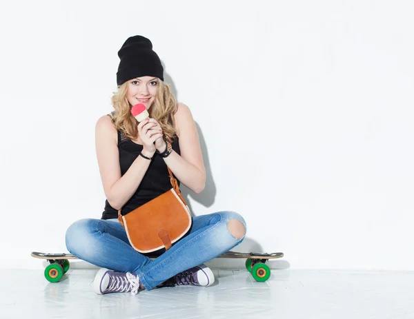 Jovem menina da moda alegre bonita em jeans, tênis, chapéu sentado em um longboard com uma bolsa vintage no ombro e comer sorvete. No interior. Estúdio . — Fotografia de Stock