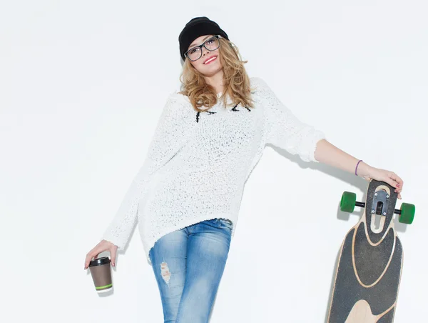 Giovane ragazza allegra alla moda in un maglione bianco con un bicchiere di caffè e longboard posa vicino alla parete bianca. In studio. — Foto Stock