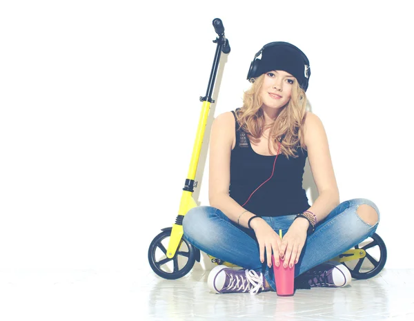 Joven hermosa chica alegre de moda en jeans, zapatillas de deporte, sombrero se sienta en un scooter amarillo y escuchar música en los auriculares. Mantiene el vidrio rosa fresco en la mano. Tonificado en colores cálidos —  Fotos de Stock