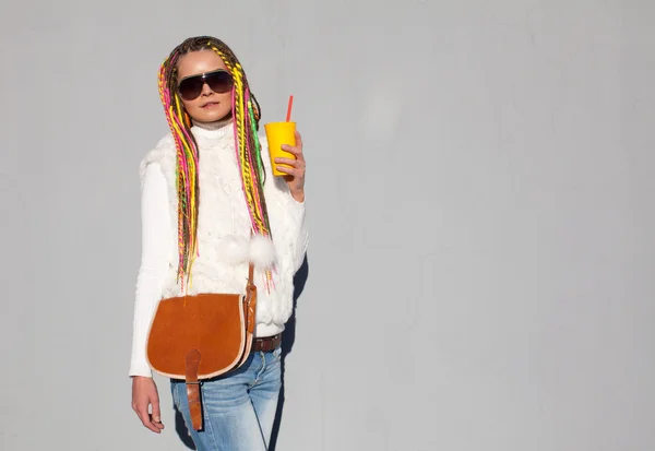 Mooi meisje met gekleurde dreadlocks zonnige zomerdag in een witte jas met zonnebril houden een gele glas met een vintage bruine tas — Stockfoto