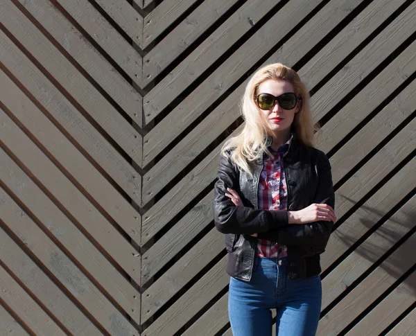 Beautiful blonde girl in huge sunglasses and a black jacket posing nex to wooden wall on a sunny day — Stock Photo, Image