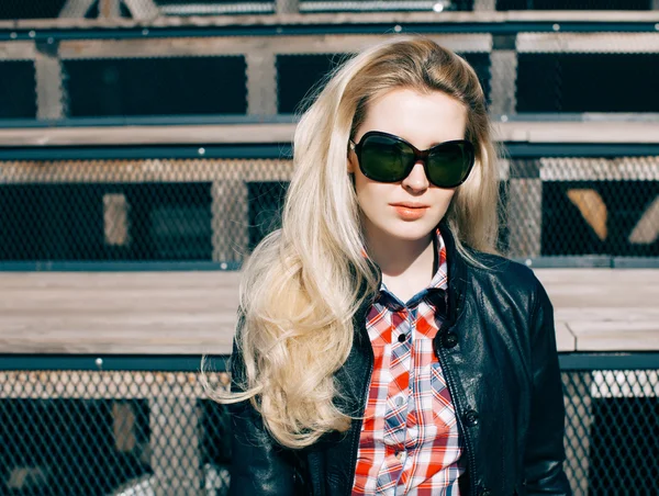 Beautiful blonde girl in huge sunglasses and a black jacket sitting on the steps of a sunny day — Stock Photo, Image