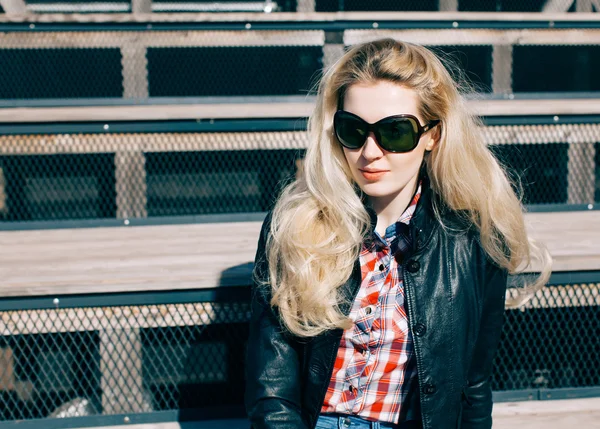 Schöne blonde Mädchen mit riesiger Sonnenbrille und schwarzer Jacke sitzt auf den Stufen eines sonnigen Tages — Stockfoto
