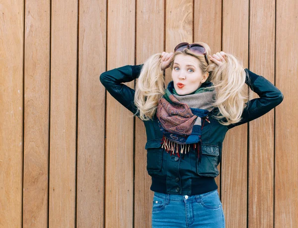 Beautiful blonde girl in black jacket posing nex to wooden wall on a sunny day and playing with her hair — Stock Photo, Image