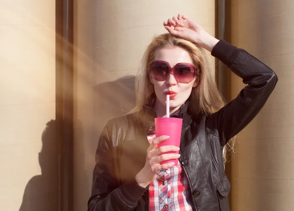Beautiful sexy blonde with sunglasses drinking beverage through a straw on a hot summer day near the wall — Stock Photo, Image