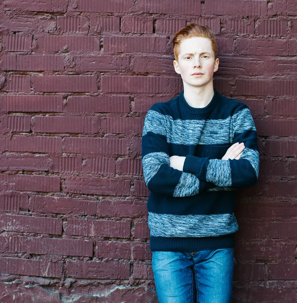 Unga rödhårig man i en tröja och jeans står bredvid en tegelvägg med korslagda armar — Stockfoto