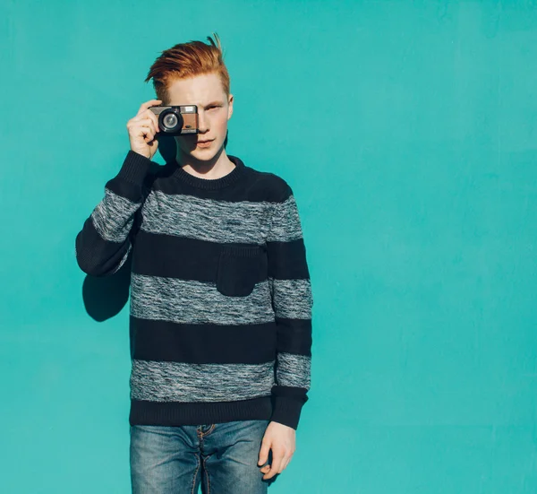 Young redhead man in a sweater and jeans standing next to turquoise wall and taking photos vintage camera warm summer sunny day — Stock Photo, Image