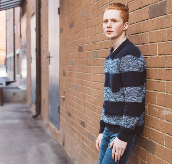 Jonge roodharige man in een trui en jeans permanent naast een oranje bakstenen muur — Stockfoto