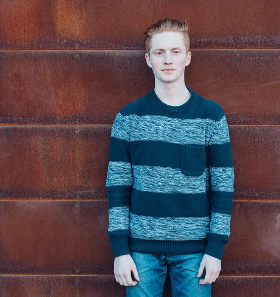 Joven pelirrojo en un suéter y jeans de pie junto a la pared oxidada de metal y sonriendo —  Fotos de Stock