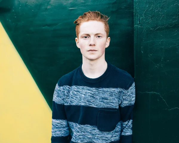 Young redhead man in a sweater and jeans standing next to green and yellow wall — Stock Photo, Image