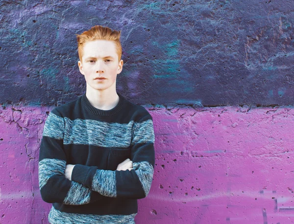 Rudy młody człowiek w sweter i dżinsy stojący obok fioletowe ściany z założonymi rękami — Zdjęcie stockowe