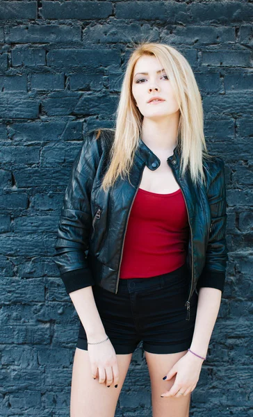 Portret van een jonge mooie blonde meisje in een zwarte jas en broek poseren in de buurt van bakstenen muur — Stockfoto