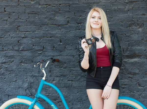 Retrato de una joven hermosa rubia con una chaqueta negra y pantalones cortos posando cerca de la pared de ladrillo junto a una bicicleta vintage azul brillante sosteniendo una cámara vintage —  Fotos de Stock