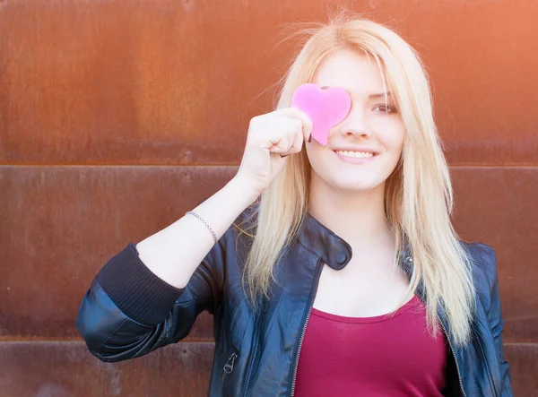 Porträtt av en vacker blond tjej med ena handen stänger ögonen rosa papper hjärtat. Filter, ljus läcka effekt. — Stockfoto