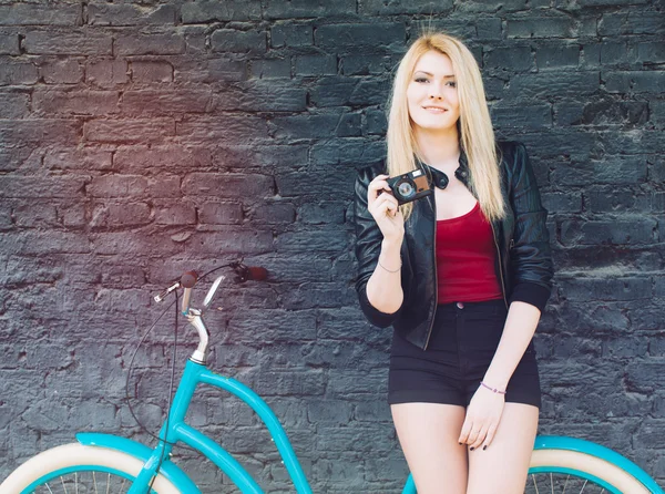 Retrato de una joven rubia hermosa con una chaqueta negra y pantalones cortos posando cerca de la pared de ladrillo junto a una bicicleta vintage azul brillante sosteniendo una cámara vintage. Filtro, efecto de fuga de luz . —  Fotos de Stock