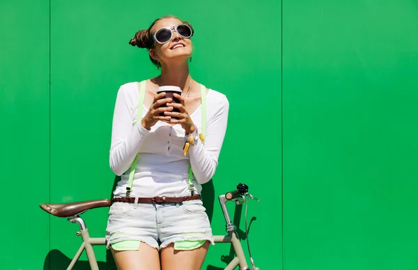 Fashionable beautiful young Pretty girl in shorts and t-shirt and cool sunglasses stands with  bicycle fix gear nex to green wall bright sunny day — Stock Photo, Image