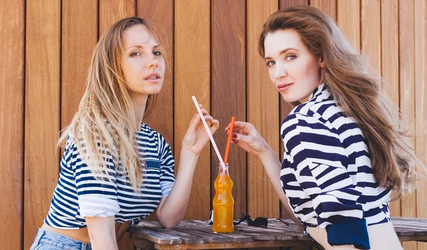 Zwei modische schöne Mädchen sitzen in einem Sommercafé und trinken orangefarbene Getränke durch einen Strohhalm aus einer Flasche. sonnig warmer Sommertag. Außenbereich. — Stockfoto