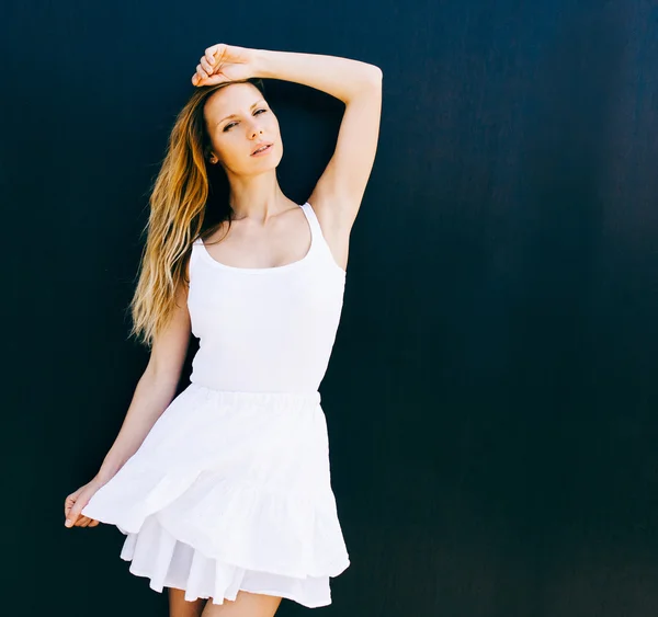 Portrait of very beautiful young blond woman in a short white dress posing on the street near a black wall. Sunny day. The wind blows her hair. She picks up the edge of the dress — Stock Fotó