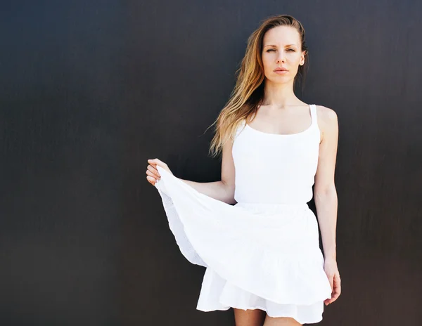 Menina loira muito bonita em um vestido branco curto posando na rua perto de uma parede preta. Dia ensolarado. O vento sopra o cabelo dela. Ela pega a borda do vestido. Retrato — Fotografia de Stock