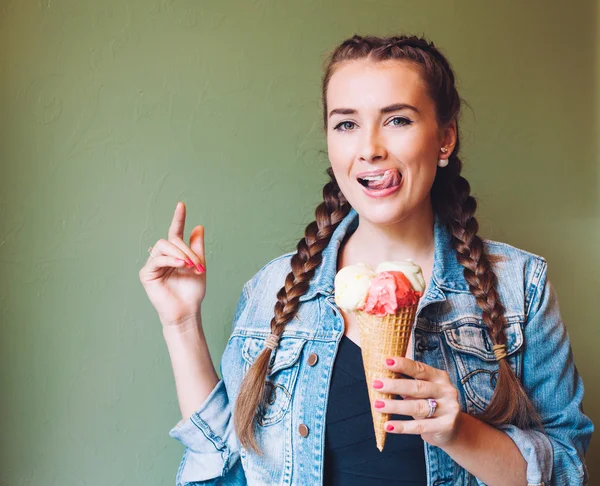 Beautiful girl with braids sitting in a cafe and eating huge multi-colored ice cream in a waffle cone. Smiling at the camera and licking lips. — Stockfoto