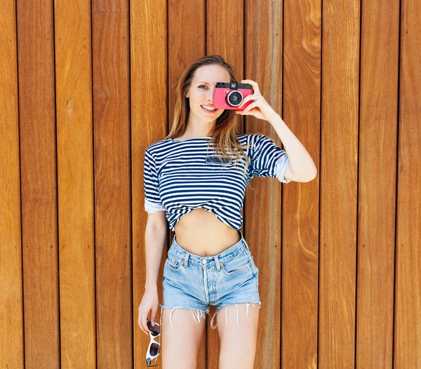 Young sexy smiling photographer woman, holding and making picture on retro pink vintage camera, wearing bright marine clothes, joy and happiness. On background wall of wooden planks Stock Fotó