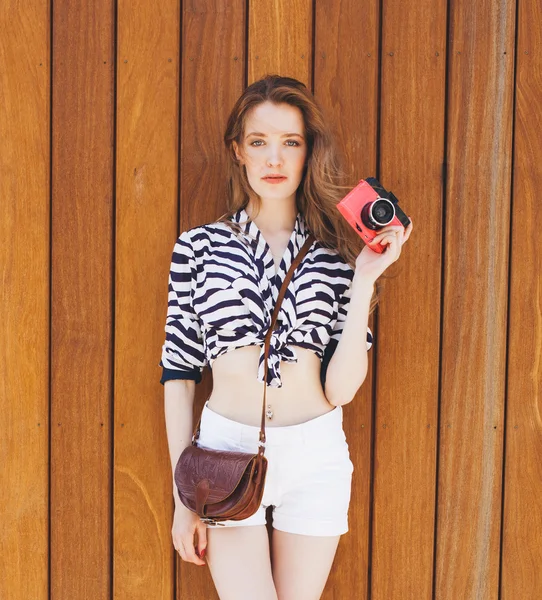 Young sexy smiling photographer woman, holding and making picture on retro ???? vintage camera, wearing bright marine clothes, with vintage bag, joy and happiness. On background wall of wooden planks Jogdíjmentes Stock Képek