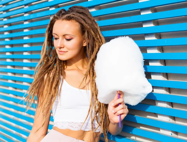 Hermosa mujer joven con rastas posando cerca de la pared de tablón azul con algodón de azúcar de verano noche caliente —  Fotos de Stock