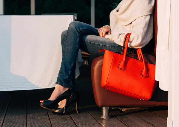 Chica sentada en el sofá de la cafetería de verano con un gran rojo super bolsos de moda en un suéter jeans y zapatillas de deporte en una cálida noche de verano. colores cálidos Fotos de stock libres de derechos