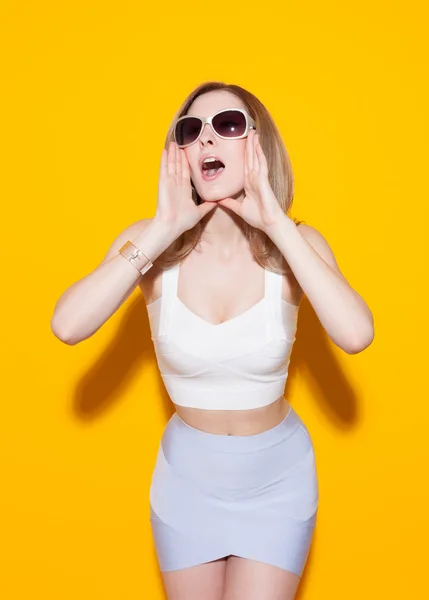 Fashionable Beautiful girl screaming with her hands folded horn on a yellow bright background. She wore a light top and short skirt and sunglasses. Gorgeous Woman Portrait. Stylish Haircut and Makeup. — Stockfoto