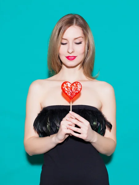 Bella ragazza alla moda che tiene un cuore di caramelle rosse e guarda il suo. In un abito nero su sfondo verde in studio. Fashion Beauty Girl. Splendido ritratto di donna. Elegante taglio di capelli e trucco. Ah! — Foto Stock