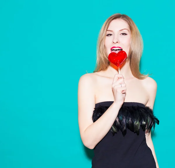 Fashionable beautiful girl biting a red lollipop and look at his. In a black dress on a green background in the studio. Look at the camera. Fashion Beauty Girl. Sexy Glamour Girl on Green background. — Stockfoto