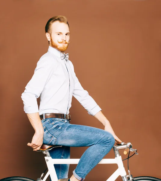 Un jeune homme à la moustache et à la barbe est assis sur un vélo moderne à la mode. Jeans et chemise, le style hipster du nœud papillon. Couleur tonique — Photo