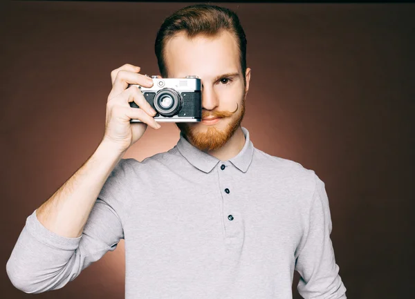 Портрет молодого человека с усами, джинсами с бородой и клетчатой рубашкой в стиле хипстера в студии, делающего крупный план винтажной камеры. Смотреть в камеру — стоковое фото
