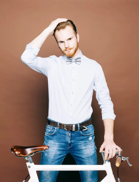 En ung man med mustasch och skägg är nära fashionabla moderna fixgear cykel. Smidig hår på huvudet. Jeans och skjorta, fluga hipster style. Tonad färg — Stockfoto