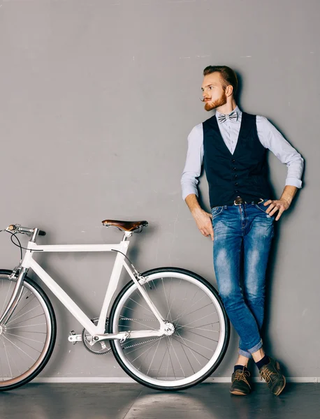 Młody człowiek z wąsy i brodę znajduje się w pobliżu modne nowoczesne fixgear rowerów. Dżinsy i koszula, kamizelka i muszka hipster stylu. Stonowanych kolorów — Zdjęcie stockowe