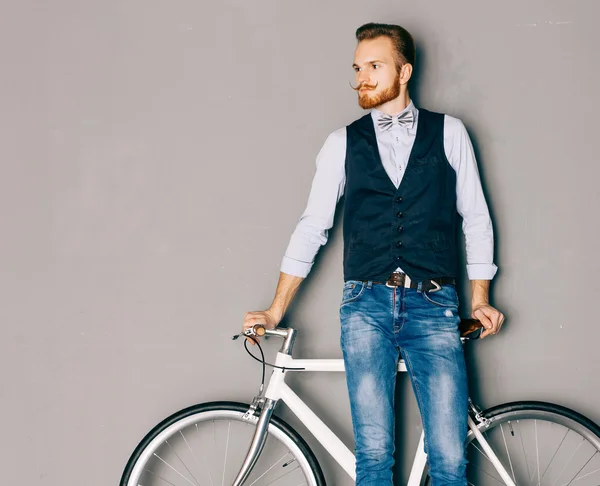 Un jeune homme avec la moustache et la barbe est à proximité du vélo fixgear moderne à la mode. Jeans et chemise, gilet et le style hipster nœud papillon. Fond gris. Couleur tonique — Photo