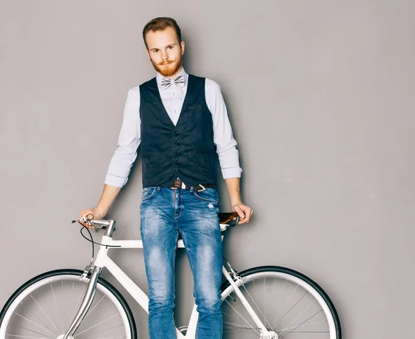 Młody człowiek z wąsy i brodę znajduje się w pobliżu modne nowoczesne fixgear rowerów. Dżinsy i koszula, kamizelka i muszka hipster stylu. Szarym tle. Stonowanych kolorów. Patrząc na kamery — Zdjęcie stockowe