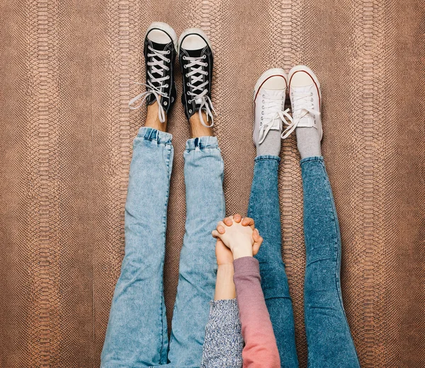 Jovens pernas de casal de moda em jeans e tênis pés para cima e de mãos dadas na parede. Fecha. Cor quente. Interior . — Fotografia de Stock