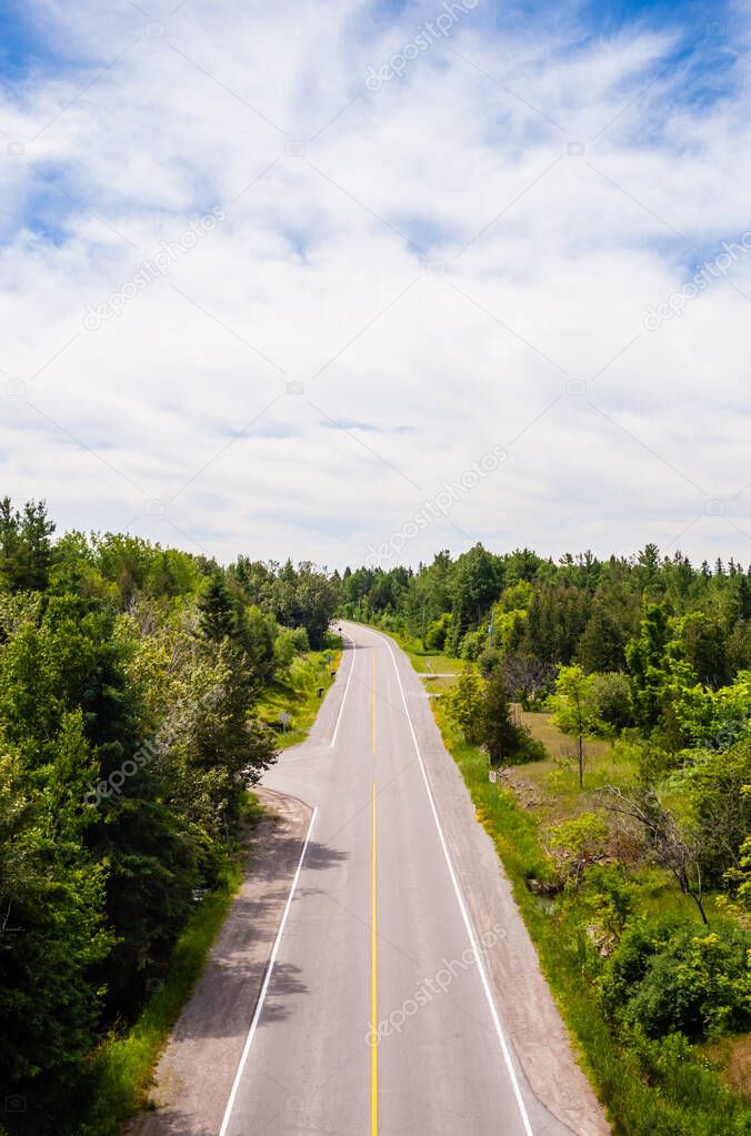 Long empty two-lane road in forested area curving left in distance under partly cloudy sky.
