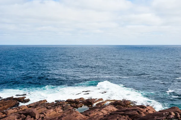 Su ve bulutlu gökyüzü altında kayalık okyanus kıyısında sıçramasına dalgalar — Stok fotoğraf