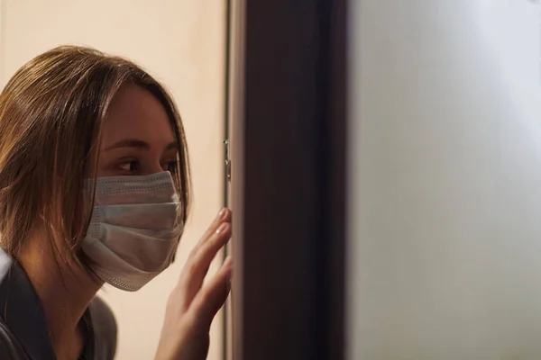 Young woman in medical mask looking through peephole of front door in apartment when somebody rings doorbell. Stay home and self isolation concept. Home quarantine, prevention COVID-19.